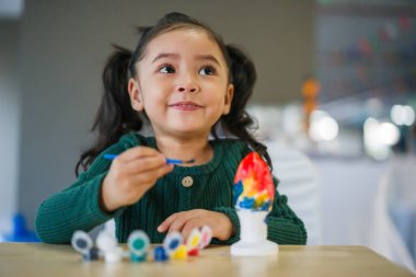 Çocuk kız masaya alçı heykel boyuyor.