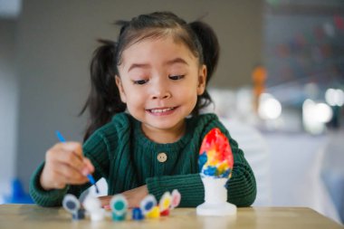 Çocuk kız masaya alçı heykel boyuyor.