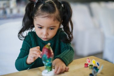 Çocuk kız masaya alçı heykel boyuyor.