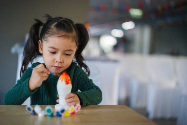 Çocuk kız masaya alçı heykel boyuyor.