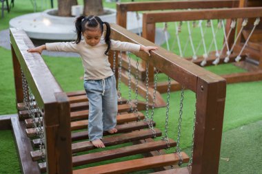 Parktaki oyun parkında oynayan ve tahta halat köprüsünde yürüyen bir çocuk.