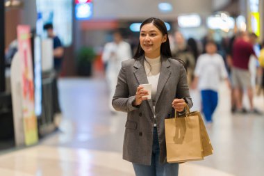 Alışveriş merkezinde alışveriş yaparken elinde bir fincan kahve ve poşet tutan bir kadın.