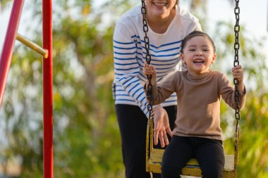 Neşeli kız ve annesi oyun parkında salıncakta oynuyorlar.