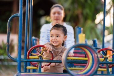Çocuk kız annesiyle oyun parkında direksiyon oyuncağıyla oynuyor.