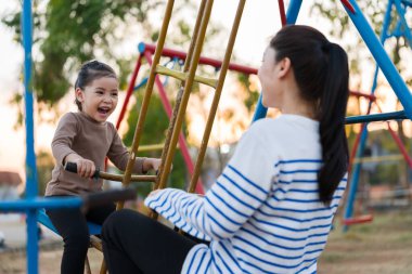 Neşeli çocuk annesi ile açık hava çocuk parkında çifte salıncakta oynuyor.