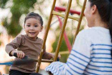 Neşeli çocuk annesi ile açık hava çocuk parkında çifte salıncakta oynuyor.