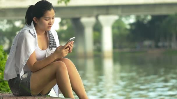 Молода азіатка використовує смартфон і виглядає як розслабляюча на відкритому повітрі, виглядає щасливою і усміхненою: повний HD — стокове відео