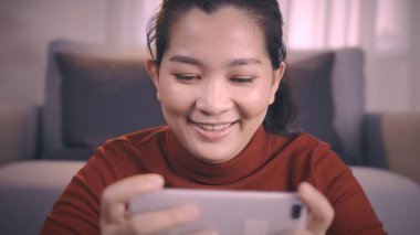 Kırmızı gömlekli Asyalı kadınlar online oyun oynamak için akıllı telefon kullanıyorlar oturma odasında heyecanlı duygular var.