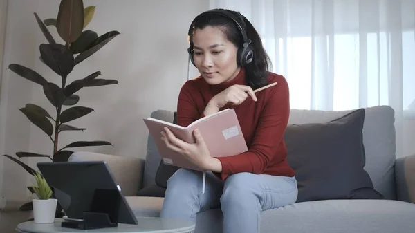 Asian woman on redshirt using a tablet with headphone for meeting online at home in coronavirus situation new normal work from home