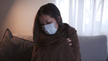 Coronavirus 'tan muzdarip genç Asyalı kadının gözleri morarmış. Üşümüş ve maske ve battaniye kullanmış. Ayrı oturma odasındaki kanepede ve karantinada kalmak için.