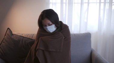 Coronavirus 'tan muzdarip genç Asyalı kadının gözleri morarmış. Üşümüş ve maske ve battaniye kullanmış. Ayrı oturma odasındaki kanepede ve karantinada kalmak için.