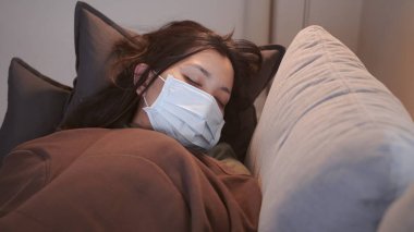 Coronavirus 'tan muzdarip genç Asyalı kadının gözleri morarmış. Üşümüş ve maske ve battaniye kullanmış. Ayrı oturma odasındaki kanepede ve karantinada kalmak için.