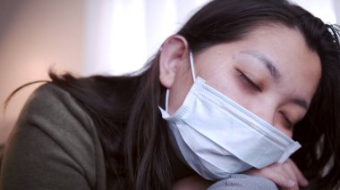 Coronavirus 'tan muzdarip genç Asyalı kadının gözleri morarmış. Üşümüş ve maske ve battaniye kullanmış. Ayrı oturma odasındaki kanepede ve karantinada kalmak için.