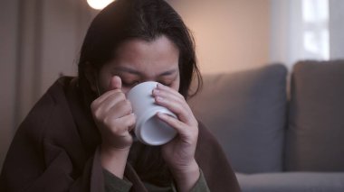 Acı çekerken ılık su içen hasta Asyalı kadının soğuk algınlığı nedeniyle tüm vücudunda soğuk göz kapakları var ve kendini ısıtmak ve karantinaya almak için maske ve battaniye kullanıyor.