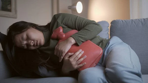 Adet sancısı çeken hasta kadın acısını dindirmek ve oturma odasındaki kanepede daha iyi hissetmek için karnına sıcak su torbası sürüyor.