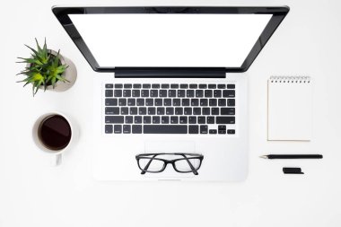 Boş ekranlı dizüstü bilgisayar bir sürü malzemeyle birlikte beyaz ofis masasının üstünde. Kopyalama alanı olan üst görünüm, düz yatay.