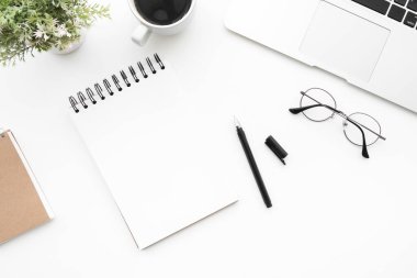 Boş defter sayfası beyaz ofis masasının üstünde dizüstü bilgisayarı, kahve ve malzemelerle birlikte. Kopyalama alanı olan üst görünüm, düz yatay.