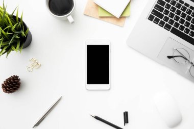 Beyaz ekranlı akıllı telefon beyaz ofisin üstünde dizüstü bilgisayar, alet edevat ve malzemelerle birlikte. Kopyalama alanı olan üst görünüm, düz yatay.