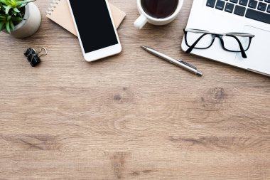 Akıllı telefonlu ahşap ofis masası. Boş ekran, dizüstü bilgisayar, kahve fincanı ve malzemeler. Kopyalama alanı olan üst görünüm, düz yatay.