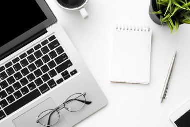 Boş sayfalı küçük bir defter, beyaz ofis masasının üstünde dizüstü bilgisayar ve malzemelerle birlikte. Üst manzara, düz uzanma.