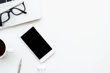 Beyaz akıllı telefon beyaz ekranlı en üst beyaz ofis masasında dizüstü bilgisayar, kahve ve malzeme ile birlikte. Kopyalama alanı olan üst görünüm, düz yatay.