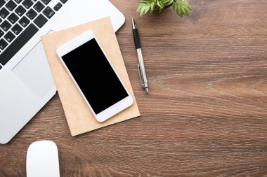 Boş maket ekranlı akıllı telefon, ahşap ofis masasının üstünde dizüstü bilgisayar ve malzemelerle birlikte. Kopyalama alanı olan üst görünüm, düz yatay.