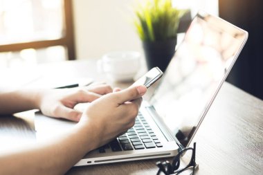 Erkek elleri, ofisteki akıllı telefon ekranına dizüstü bilgisayar ve malzemelerle dokunuyor..
