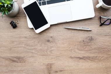 Akıllı telefon ahşap ofis masasının üstünde dizüstü bilgisayar, kahve ve malzemelerle birlikte. Kopyalama alanı olan üst görünüm, düz yatay.