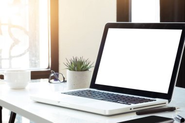 Boş maket ekranı olan dizüstü bilgisayar kahve ve malzemelerle dolu beyaz ofis masasının üstünde..