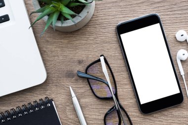 Boş ekranlı akıllı telefon ahşap ofis masasının üstünde dizüstü bilgisayar ve ofis malzemeleriyle birlikte. Üst manzara, düz uzanma.