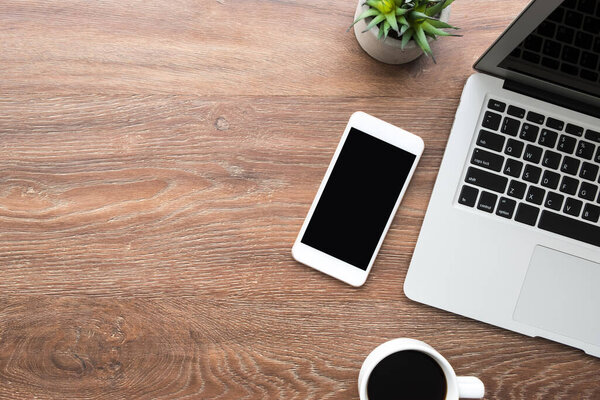 Wood office desk table with white smartphone with blank mock up screen, laptop computer, cup of coffee and supplies. Top view with copy space, flat lay.