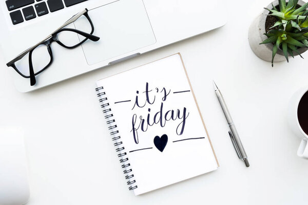 Notebook with it's friday text written on it on white office desktable with laptop computer and other office supplies. Top view with, flat lay, tgif concept.