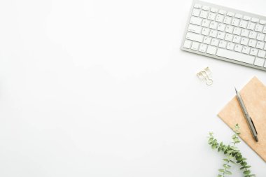 Bilgisayar klavyeli beyaz ofis masası, kalem ve malzemelerle dolu defter. Kopyalama alanı olan üst görünüm, düz yatay.