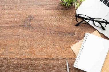 Ahşap ofis masasında boş defter, kalem, dizüstü bilgisayar ve malzemeler. Kopyalama alanı olan üst görünüm, düz yatay.