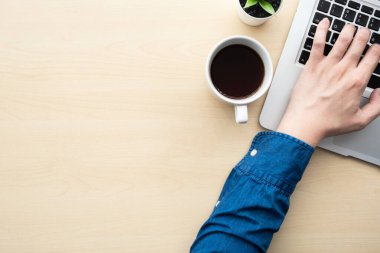 Erkek eli, bir fincan kahveyle ahşap ofis masasının üzerinde dizüstü bilgisayarda klavye kullanıyor. Kopyalama alanı olan üst görünüm, düz yatay.