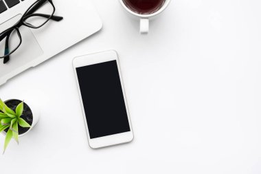Boş ekranlı akıllı telefonlu beyaz ofis masası, dizüstü bilgisayar, bir fincan kahve ve malzeme. Kopyalama alanı olan üst görünüm, düz yatay.