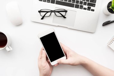 Adam bilgisayar ve malzemelerle beyaz ofis masasının üzerinde boş ekranlı akıllı telefon kullanıyor. Üst görünüm.