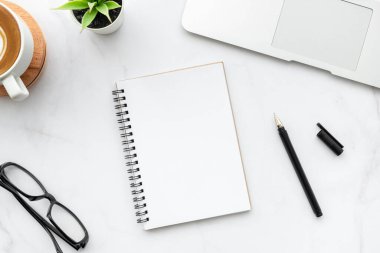 Kalemli boş defter, beyaz mermer ofis masasının üstünde dizüstü bilgisayarı, kahve ve malzemelerle dolu. Üst manzara, düz uzanma.
