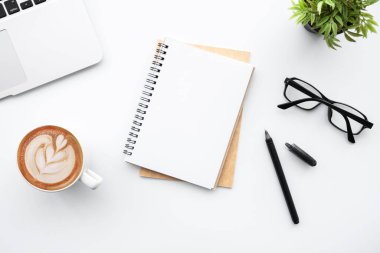 Beyaz ofis masasının üstünde dizüstü bilgisayarı, kahve ve malzemelerle dolu boş bir defter var. Kopyalama alanı olan üst görünüm, düz yatay.