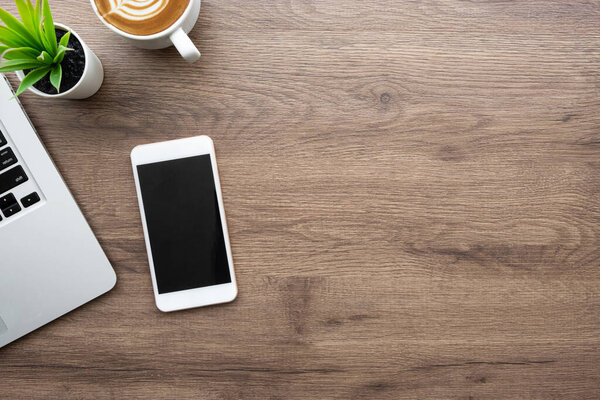 Wood office desk table with smartphone with blank screen, laptop computer, cup of coffee and supplies. Top view with copy space, flat lay.