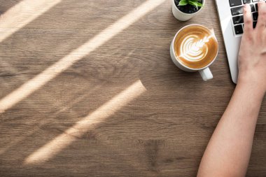 Adam ahşap ofis masasının üzerinde bir fincan kahveyle dizüstü bilgisayarda tuşlara basıyor. Sabah konseptiyle evden çalışmak.