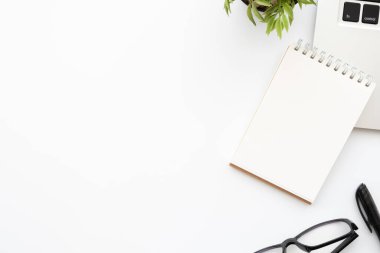 Küçük boş defter, dizüstü bilgisayar ve ofis malzemeleri olan beyaz ofis masası. Kopyalama alanı olan üst görünüm, düz yatay.