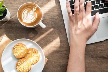 Adam, ahşap ofis masasının üzerinde bir fincan kahve ve bir tabak kurabiyeyle dizüstü bilgisayarda tuş basıyor. Sabah konseptiyle evden çalışmak.