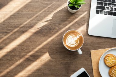 Dizüstü bilgisayarlı ahşap ofis masası, bir tabak bademli kurabiye ve sabah güneşli bir fincan latte kahve. Ev konsepti üzerinde çalışıyorum. Kopyalama alanı olan üst görünüm, düz yatay.