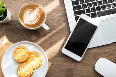 Beyaz akıllı telefon dizüstü bilgisayarın üstünde, ahşap ofis masasının üzerinde bir tabak kurabiye, bir fincan kahve ve malzemeyle birlikte. Top view, evden çalışmak sabah konsepti..