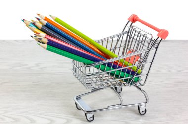 Minyatür bir alışveriş arabasının içindeki renkli kalemler beyaz arka planda beyaz ahşap bir masada.