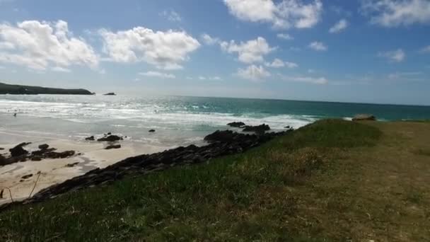 Una sartén en movimiento a lo largo de un promontorio en Newquay con vista a la playa de Fistral Bay, marea y surf con surfistas lejanos . — Vídeo de stock