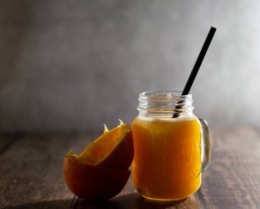 Taze portakal suyu bardakta, ahşap bir masada siyah bir tüp, arkasında gri bir duvar. Camın yanında portakal kabuğu var. Düşük tuşlu yatay fotoğraf