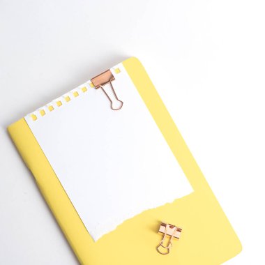 Sarı bir not defteri ve iki bakır ataç beyaz arka planda, üst tarafta. Kırtasiye ve minimalizm
