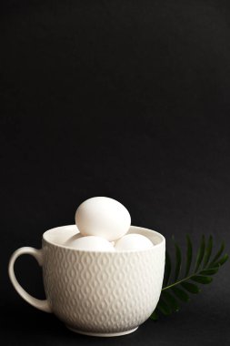 Siyah arka planda büyük beyaz bir fincanda yumurtalar, tabağın yanında bir palmiye dalı var. Minimalizm ve Paskalya Bayramı konsepti.